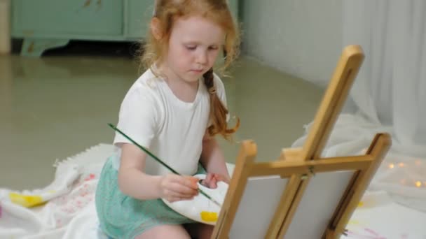 Красива руда дівчина сидить на підлозі в студії перед мольбертом і малює пензлем і кольоровими фарбами — стокове відео