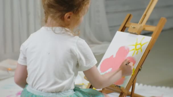 Bella ragazza dai capelli rossi si siede sul pavimento in studio di fronte al cavalletto e disegna un pennello e vernici colorate — Video Stock