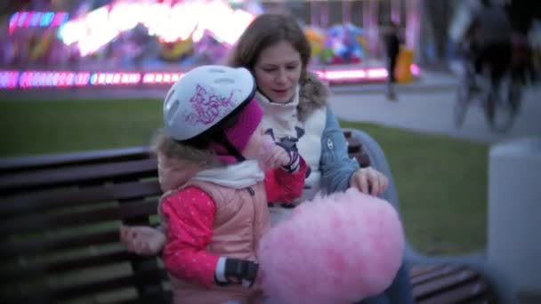 Vacker liten flicka sitter på en bänk i en nöjespark, äta rosa söt sockervadd rullande på rullskridskor — Stockvideo