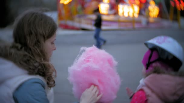 Menina bonita sentada em um banco em um parque de diversões, comendo doces de algodão doce rosa rolando em patins — Vídeo de Stock