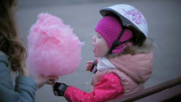 Красивая маленькая девочка сидит на скамейке в парке аттракционов и ест розовую сладкую сладкую вату, катающуюся на роликовых коньках — стоковое видео