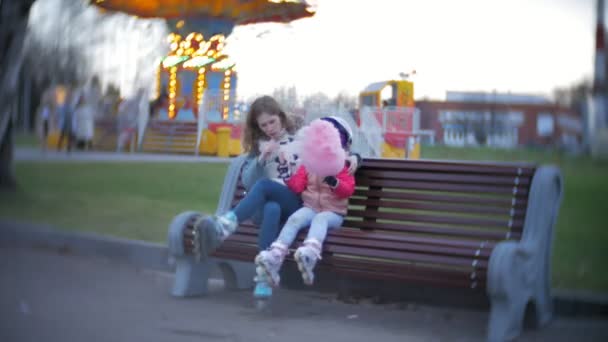 Belle petite fille assise sur un banc dans un parc d'attractions, mangeant de la barbe à papa rose roulante sur des patins à roulettes — Video