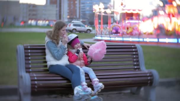 Hermosa niña sentada en un banco en un parque de atracciones, comiendo caramelos rosados de algodón dulce rodando sobre patines — Vídeo de stock
