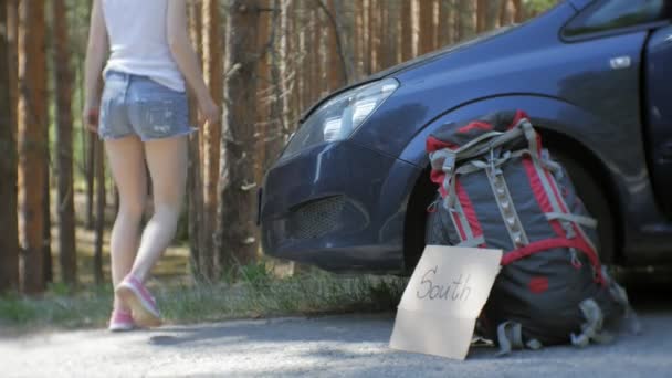 Junge schöne Frau trampt auf der Straße stehend mit einem Rucksack auf einem Tisch mit der Aufschrift Süden in der Nähe eines kaputten Autos — Stockvideo