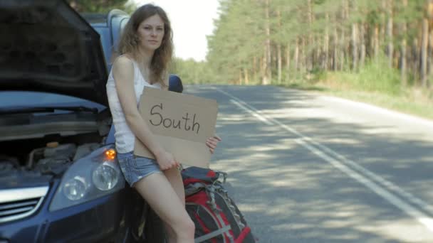 Giovane bella donna autostop in piedi sulla strada con uno zaino su un tavolo con un'iscrizione SUD vicino a un'auto rotta — Video Stock