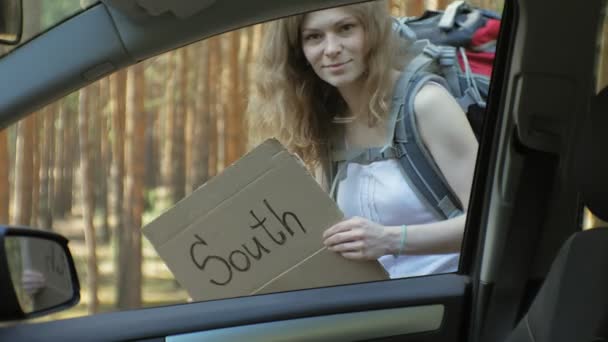 Jeune belle femme auto stop debout sur la route avec un sac à dos sur une table avec une inscription SUD près d'une voiture cassée — Video