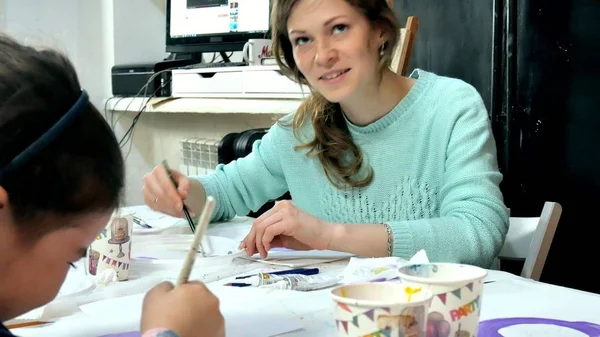 Bambini ragazzi e ragazze seduti insieme intorno al tavolo in classe e disegno. Con loro c'è la loro giovane e bella maestra . — Foto Stock