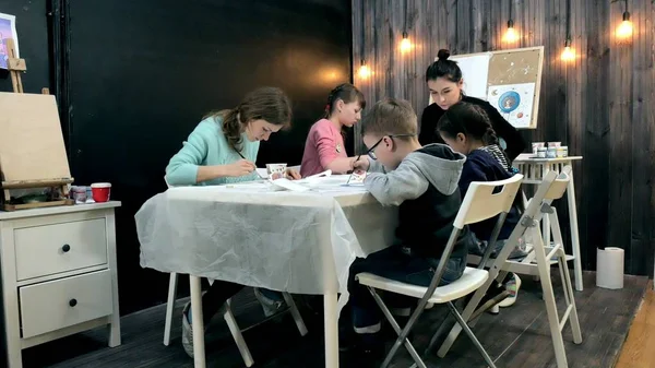 Bambini ragazzi e ragazze seduti insieme intorno al tavolo in classe e disegno. Con loro c'è la loro giovane e bella maestra . — Foto Stock