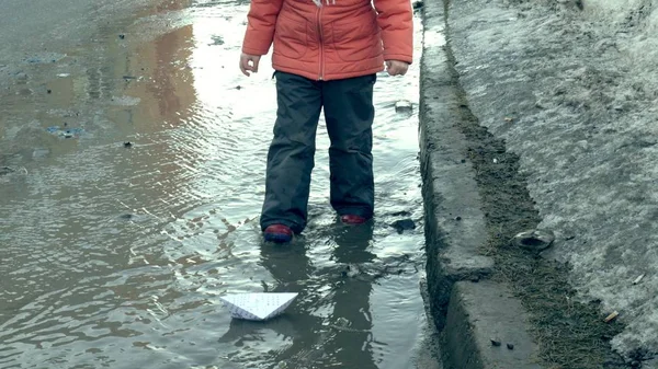 Girl in rubber boots jumping on puddle near wite paper boat — Stock Photo, Image