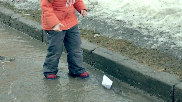 Mädchen in Gummistiefeln springt auf Pfütze nahe weißem Papierboot — Stockfoto