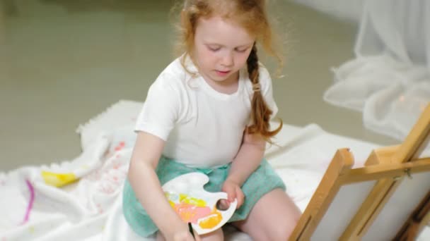 Красива руда дівчина сидить на підлозі в студії перед мольбертом і малює пензлем і кольоровими фарбами — стокове відео