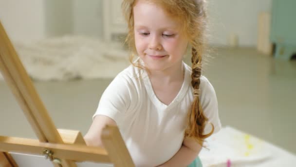 Beautiful red-haired girl sits on the floor in the studio in front of the easel and draws a brush and colored paints — Stock Video