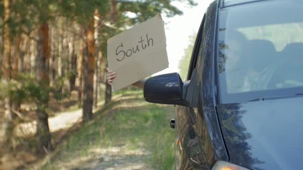 Молодая красивая женщина автостопом стоит на дороге с рюкзаком на столе с надписью Юг в машине — стоковое видео