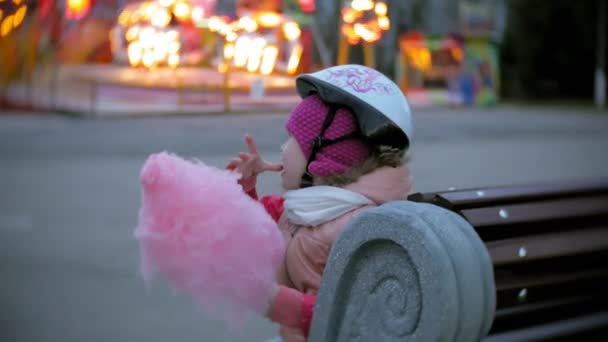 Hermosa niña sentada en un banco en un parque de atracciones, comiendo caramelos rosados de algodón dulce rodando sobre patines — Vídeos de Stock