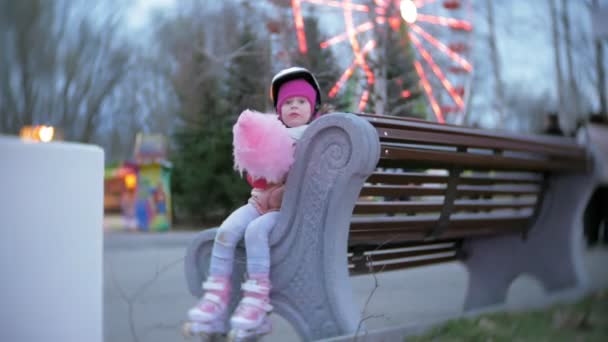 Красива маленька дівчинка сидить на лавці в парку розваг, їсть рожеві солодкі бавовняні цукерки, що катаються на роликах — стокове відео