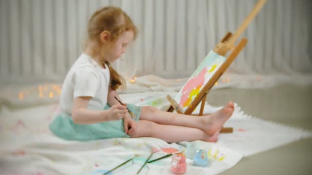 Hermosa chica pelirroja se sienta en el suelo en el estudio delante del caballete y dibuja un pincel y pinturas de colores — Vídeos de Stock