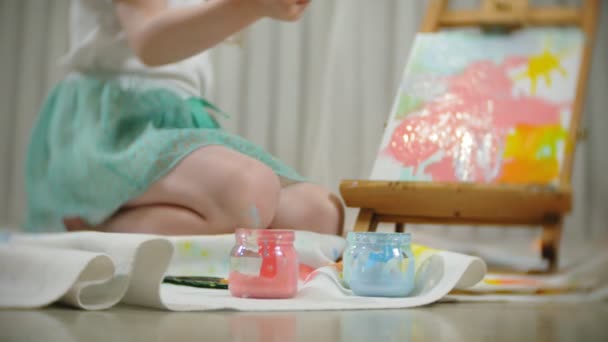 Belle fille aux cheveux roux s'assoit sur le sol dans le studio devant le chevalet et dessine un pinceau et des peintures colorées — Video