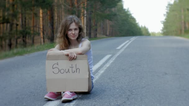 Mulher bonita jovem carona em pé na estrada com uma mochila em uma mesa com uma inscrição SOUTH — Vídeo de Stock