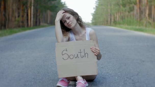 Mulher bonita jovem carona em pé na estrada com uma mochila em uma mesa com uma inscrição SOUTH — Vídeo de Stock