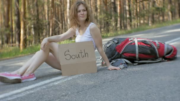 Mulher bonita jovem carona em pé na estrada com uma mochila em uma mesa com uma inscrição SOUTH — Vídeo de Stock