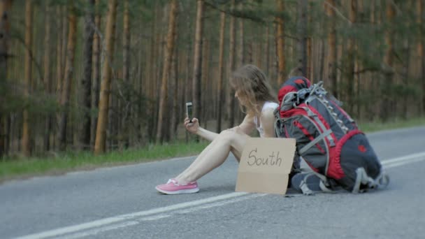 Νεαρή όμορφη γυναίκα ωτοστόπ στέκεται στο δρόμο με ένα σακίδιο σε ένα τραπέζι με μια επιγραφή Νότια — Αρχείο Βίντεο