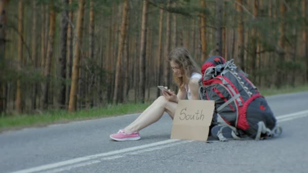 Giovane bella donna autostop in piedi sulla strada con uno zaino su un tavolo con un'iscrizione SUD — Video Stock