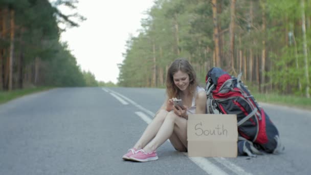 Νεαρή όμορφη γυναίκα ωτοστόπ στέκεται στο δρόμο με ένα σακίδιο σε ένα τραπέζι με μια επιγραφή Νότια — Αρχείο Βίντεο