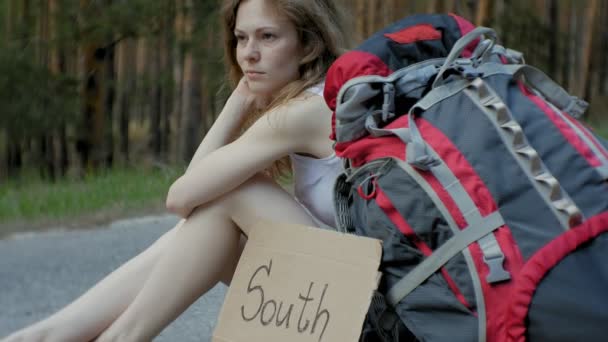 Young beautiful woman hitchhiking standing on the road with a backpack on a table with an inscription SOUTH — Stock Video