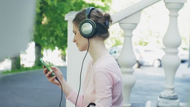 Junge, stylische, schöne Frau, die auf den Straßen der Stadt Musik mit Kopfhörern am großen Monitor hört. — Stockvideo