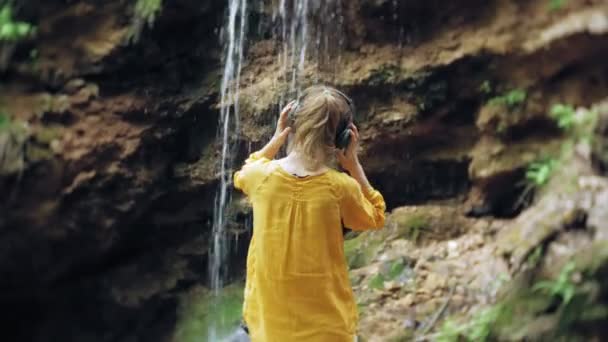 Young stylish beautiful woman, near a waterfall listening to music in large monitor headphones. — Stock Video