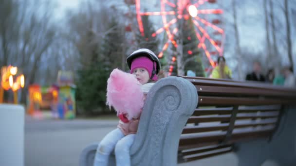 美丽的小女孩坐在一个游乐园的长凳上, 吃粉红色的甜棉花糖滚动轮滑鞋 — 图库视频影像