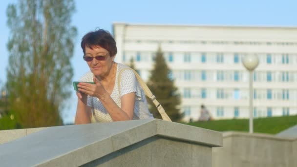 Χαμογελώντας πληκτρολόγησης μήνυμα τηλέφωνο ηλικιωμένη γυναίκα στο πάρκο. Ανώτερος γυναίκα χρησιμοποιώντας ασύρματη σύνδεση στο internet στο smart phone. Ευτυχισμένος παλιά γυναίκα κοιτάζει την οθόνη σε smartphone. — Αρχείο Βίντεο