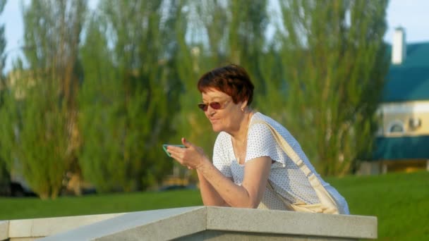 Femme âgée souriante tapant un message téléphonique au parc. Femme âgée utilisant une connexion Internet sans fil sur un téléphone intelligent. Vieille femme heureuse regardant l'écran sur smartphone . — Video