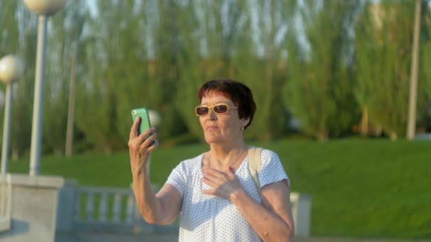 Усміхаючись літня жінка, друкує повідомлення по телефону в парку. Старша жінка, яка використовує бездротове підключення до Інтернету на смартфоні. Щаслива стара жінка дивиться на екран на смартфоні . — стокове відео