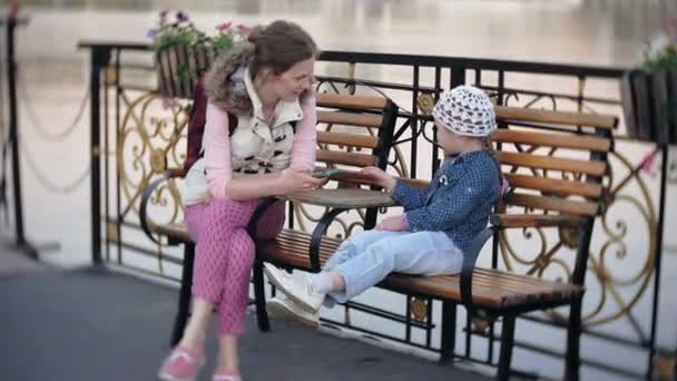 Piccola, bella, lgirl e sua madre sedute sulla panchina del parco, in mano al suo quaderno. Indossa un cappotto. Prende appunti e sembra molto romantica. . — Video Stock