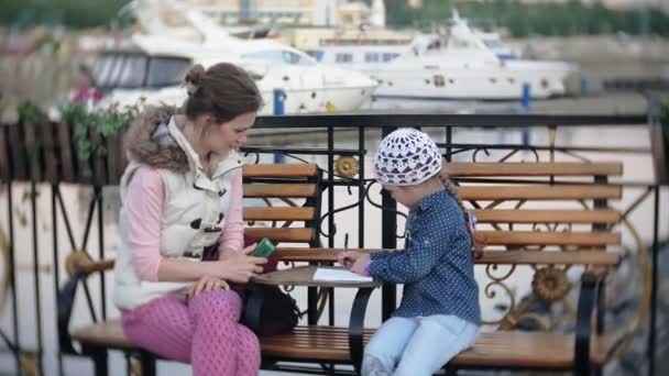 Pequeña, hermosa, niña y su madre sentadas en el banco en el parque, en manos de su cuaderno. Lleva un abrigo. Ella toma notas y se ve muy romántica . — Vídeos de Stock