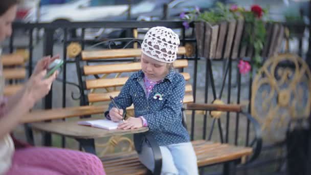 Little, beautiful, lgirl and her mother sitting on bench in park, in hands of her notebook. She is wearing a coat. She makes notes and looks very romantic. — Stock Video