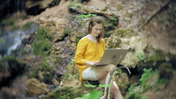 Девушка с ноутбуком, сидящая на скале, в лесу — стоковое видео