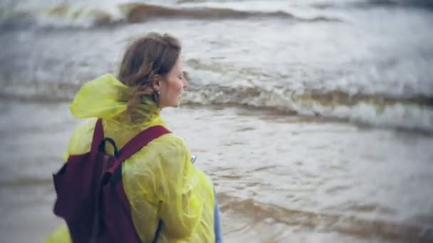 Sahil boyunca seyahat yaşam tarzı macera tatil açık havada yürüyen mutlu kadın. Bir moda sarı yağmurluk giymiş bir kız — Stok video