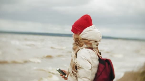 Mujer joven relajada en un sombrero rojo escuchando música y caminando a lo largo de la orilla del río. Seguimiento de la configuración en tiempo real de la toma — Vídeos de Stock