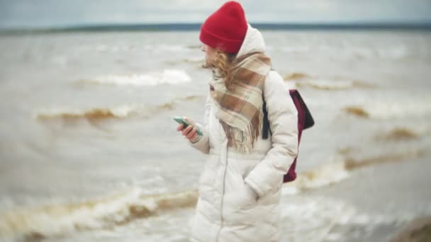 Mujer joven relajada en un sombrero rojo escuchando música y caminando a lo largo de la orilla del río. Seguimiento de la configuración en tiempo real de la toma — Vídeos de Stock