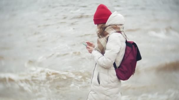 Avslappnad ung kvinna i en röd hatt lyssnar på musik och promenader längs flodstranden. Spårning i realtid inställning skottet — Stockvideo
