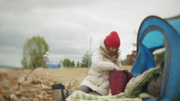 Туристическая палатка на берегу реки. две девочки сидят возле палатки, едят и пьют, смотрят на реку . — стоковое видео