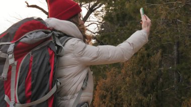 Çekici bir genç kadın kırmızı şapkalı bir büyük turistik sırt çantası ile erken baharda ormanda yürür ve telefonda fotoğraf çeker