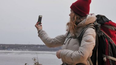 Çekici bir genç kadın kırmızı şapkalı bir büyük turistik sırt çantası ile erken baharda ormanda yürür ve telefonda fotoğraf çeker