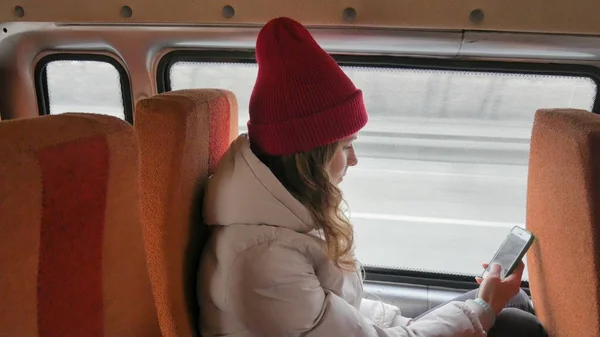 Vrolijke jongedame in een rode hoed, reizen met de bus op een trieste dag. Ze neemt foto's op een smartphone — Stockfoto