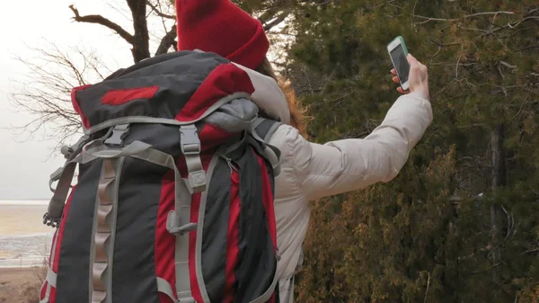 빨간 모자에는 매력적인 젊은 여자 대형 관광 배낭 이른 봄에 있는 숲을 안내 하 고 휴대 전화에서 사진을 걸립니다. — 스톡 사진