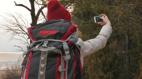 빨간 모자에는 매력적인 젊은 여자 대형 관광 배낭 이른 봄에 있는 숲을 안내 하 고 휴대 전화에서 사진을 걸립니다. — 스톡 사진