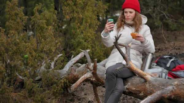 Μια ελκυστική νεαρή γυναίκα σε ένα κόκκινο καπέλο βόλτες μέσα στο δάσος, στην αρχή της άνοιξης με ένα μεγάλο τουριστικό σακίδιο, ποτά τσαγιού από ένα θερμός — Φωτογραφία Αρχείου