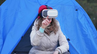 Kırmızı bir şapka çekici genç kadın turist turist çadırda oturur ve bir sanal gerçeklik kask sahiptir
